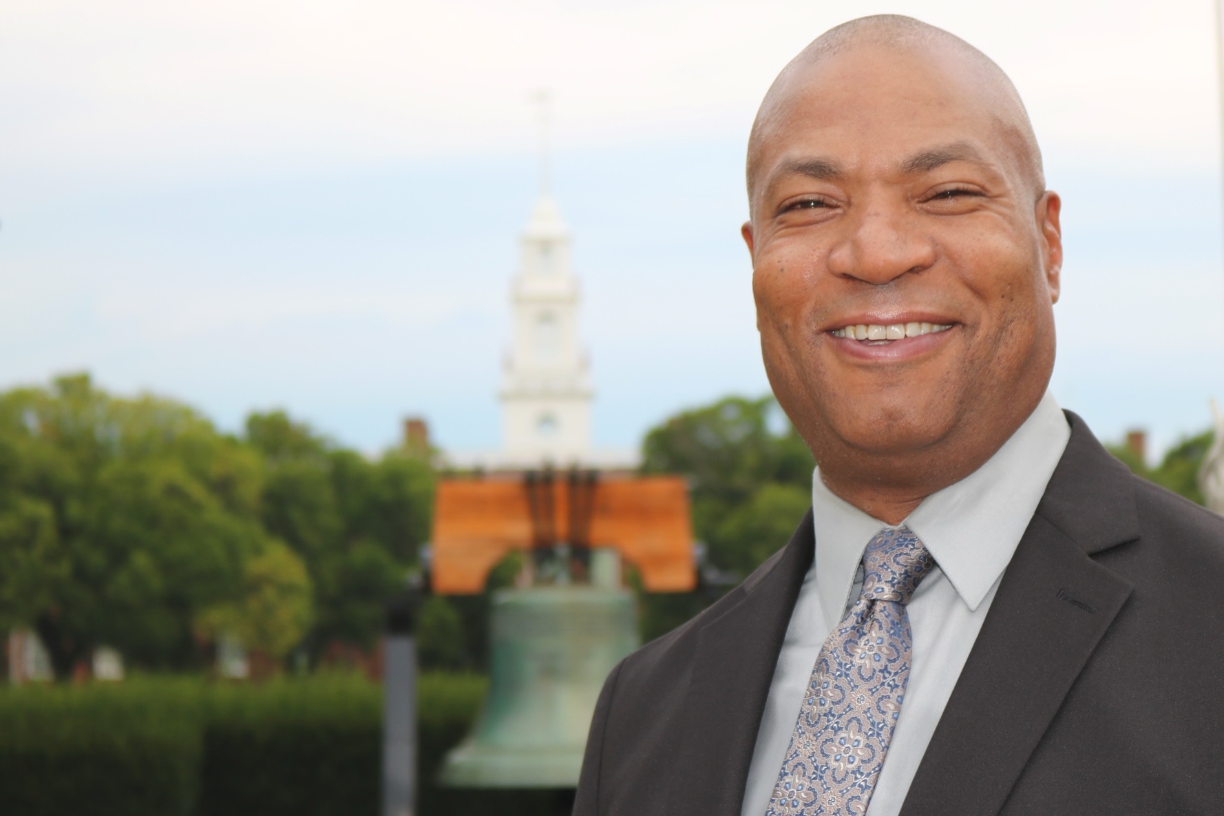 Anton Brogden smiling outside the Old State House in Dover, Delaware, with the Liberty Bell replica behind him.